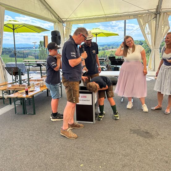 Mehrere Personen versammeln sich unter einem Zelt, wobei eine Person in ein Mikrofon spricht. Ein junger Junge beugt sich über eine Box. Eine Frau und ein weiterer Junge stehen in der Nähe.