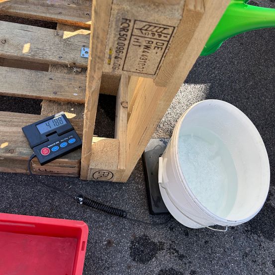 Ein Holzpalette mit einer digitalen Waage und einem weißen Eimer, der mit Wasser gefüllt ist. Die Waage zeigt 4188 Gramm an. Ein roter Behälter ist in der Nähe.