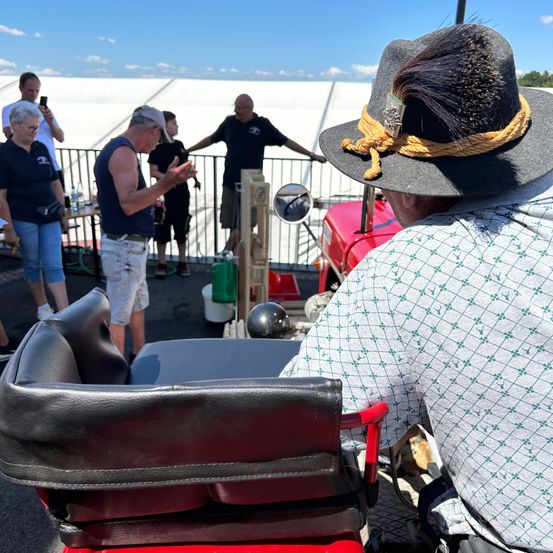 Ein Mann mit einem breitkrempigen Hut sitzt auf einem roten Stuhl draußen, während eine Gruppe von Leuten hinter einem Geländer steht.