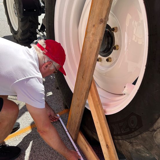 Ein Mann kniet sich hin und misst den Reifen eines Traktors mit einem Maßband, trägt eine rote Kappe und ein weißes Hemd.