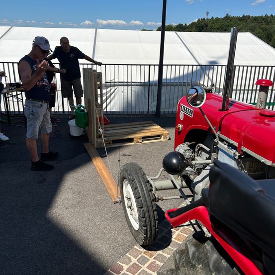 Zwei Männer stehen in der Nähe eines roten Traktors; einer gestikuliert, während der andere einen Eimer hält. Ein Holzpalette und ein Zaun sind in der Nähe. Die Szene ist im Freien, mit einem großen Zelt im Hintergrund.