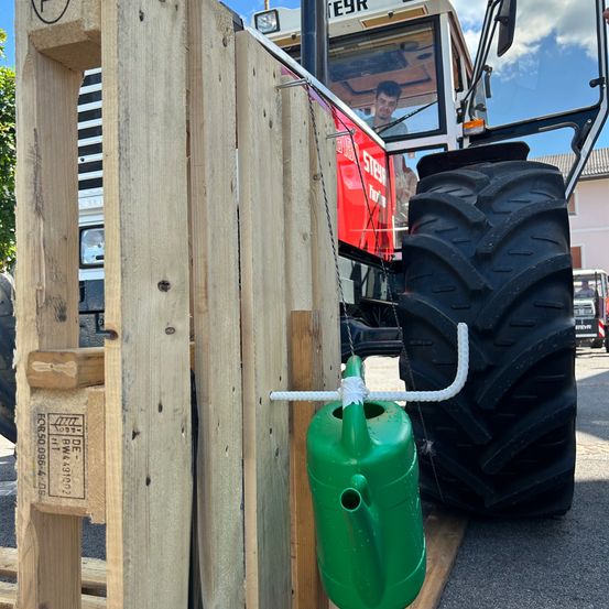 Ein Traktor mit einem großen schwarzen Reifen und einem grünen Gießkannen, der an einem Holztor hängt. Ein Mann fährt den Traktor.