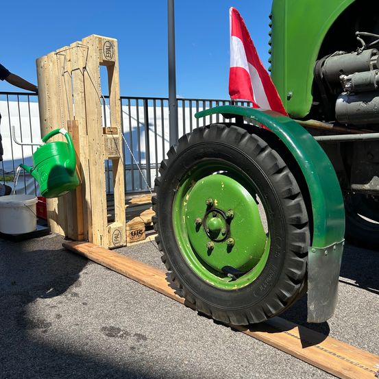 Ein grüner Gießkanne ist an einer hölzernen Struktur neben einem grünen Traktor befestigt. Ein weißer Eimer ist auf dem Boden in der Nähe der Struktur platziert.