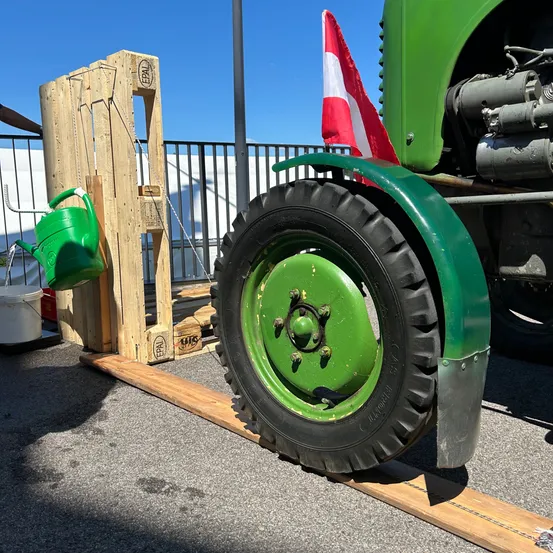 Ein grüner Traktor steht neben einem Holzständer mit einem Gießkannen und einer Flagge. Der Traktor hat ein grünes Rad und einen schwarzen Reifen.