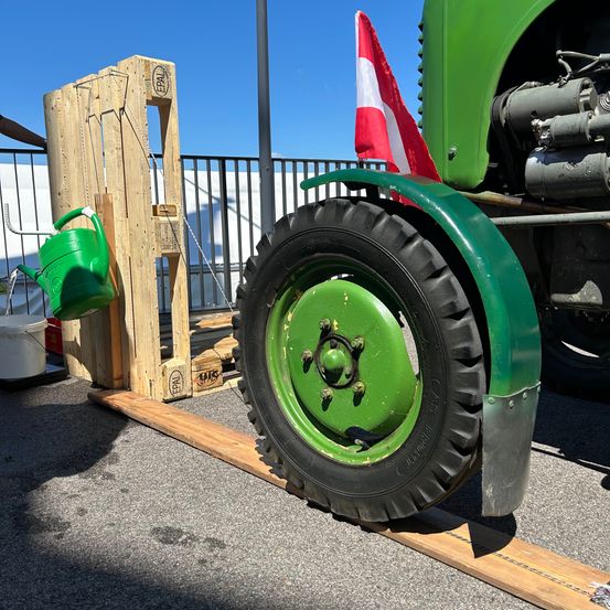 Ein grüner Traktor steht neben einem Holzständer mit einem Gießkannen und einer Flagge. Der Traktor hat ein grünes Rad und einen schwarzen Reifen.