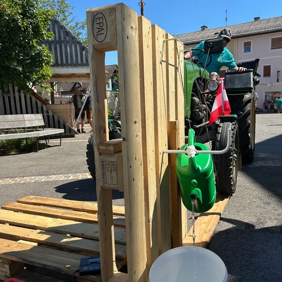 Ein Traktor mit einem grünen Gießkanister ist auf einem Holzpalette geparkt. Ein Eimer steht unter dem Gießkanister. Zwei Personen stehen in der Nähe einer Holzbank. Gebäude und Bäume sind im Hintergrund.