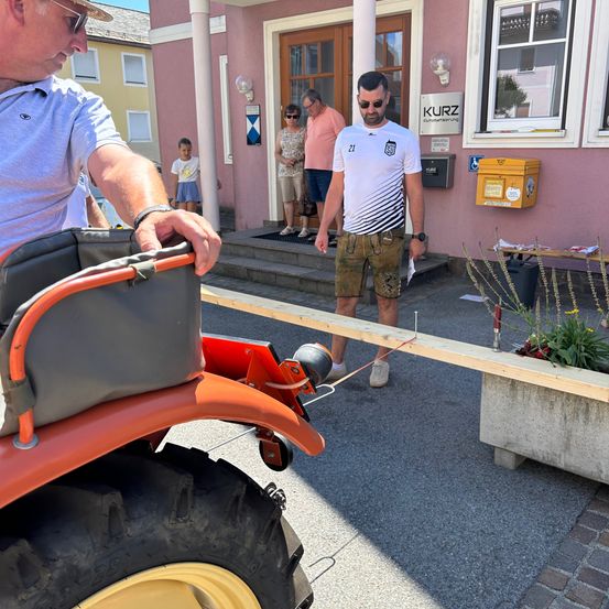 Ein Mann zieht einen roten Traktor mit einem langen orangefarbenen Rohr vor einem rosa Gebäude mit Kurz-Schild und einigen Leuten, die auf den Stufen stehen.