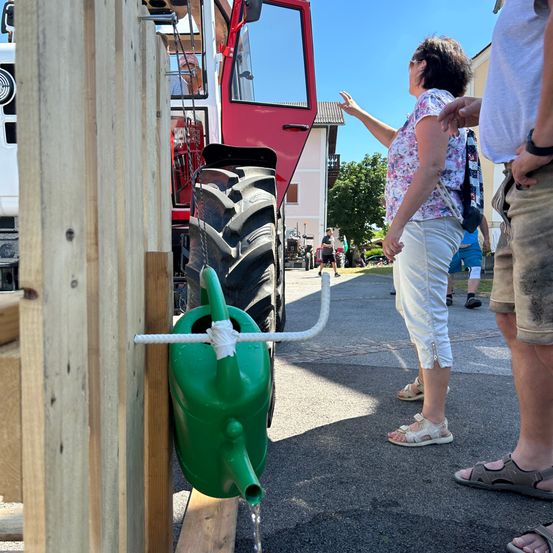 Eine Frau zeigt auf einen grünen Gießkannen, der an einem Holzpfahl neben einem roten Traktor befestigt ist, während eine weitere Person neben ihr steht. Im Hintergrund sind Menschen zu sehen, die auf der Straße gehen.