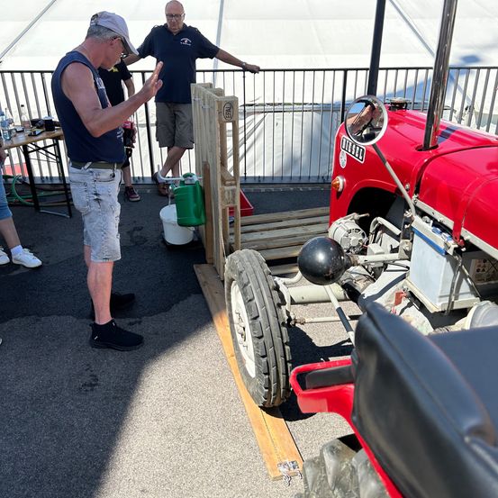 Ein Mann gestikuliert neben einem roten Traktor mit einer hölzernen Rampe, während ein anderer Mann sich auf einer Brüstung lehnt.