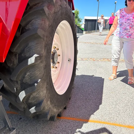Eine Frau geht an einem großen roten Traktor mit einem weißen Reifen auf einer gepflasterten Straße vorbei. Eine andere Frau geht im Hintergrund in der Nähe eines Zauns.