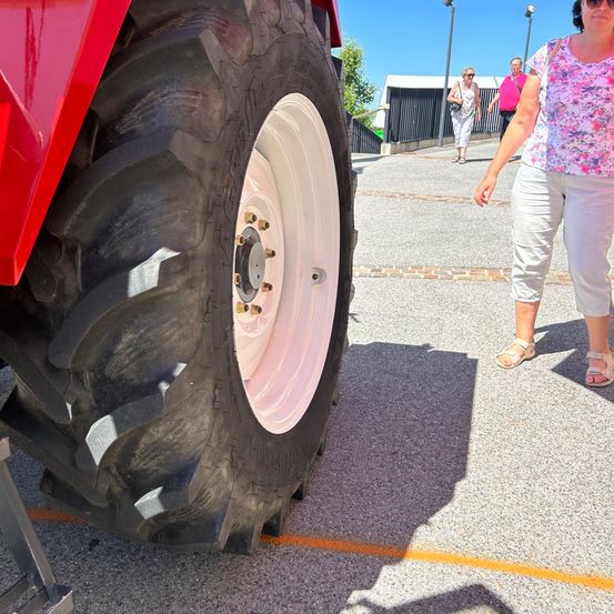 Eine Frau geht an einem großen roten Traktor mit einem weißen Reifen auf einer gepflasterten Straße vorbei. Eine andere Frau geht im Hintergrund in der Nähe eines Zauns.