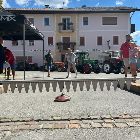 Eine Gruppe von Menschen steht vor einem Gebäude, vor dem Traktoren geparkt sind. Ein Mann wirft ein Objekt auf ein Ziel am Boden.