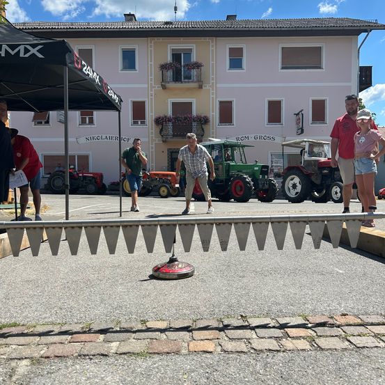 Eine Gruppe von Menschen steht vor einem Gebäude, vor dem Traktoren geparkt sind. Ein Mann wirft ein Objekt auf ein Ziel am Boden.