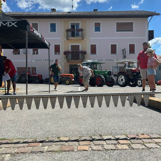 Ein Mann wirft eine Münze bei einem Münzwurfspiel. Mehrere Menschen beobachten von hinter einer Barriere aus. Dahinter sind mehrere Traktoren vor einem Gebäude geparkt.