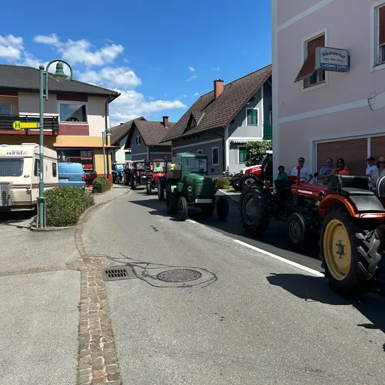 Eine Reihe von Traktoren ist am Straßenrand geparkt, wobei einige von ihnen von Menschen besetzt sind. Die Straße ist von Gebäuden gesäumt, und eine Straßenlaterne steht links. Der Himmel ist blau mit Wolken.