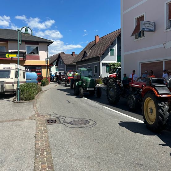 Eine Reihe von Traktoren ist am Straßenrand geparkt, wobei einige von ihnen von Menschen besetzt sind. Die Straße ist von Gebäuden gesäumt, und eine Straßenlaterne steht links. Der Himmel ist blau mit Wolken.