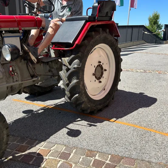 Ein Mann in Shorts sitzt auf einem roten Traktor mit schwarzen Reifen und einer weißen Felge, geparkt auf einer asphaltierten Straße.