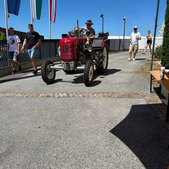 Ein Mann fährt einen roten Traktor auf einem gepflasterten Bereich. Zwei Männer gehen hinter ihm. Eine Frau geht rechts, in der Nähe eines weißen Zauns. Flaggen sind im Hintergrund zu sehen.