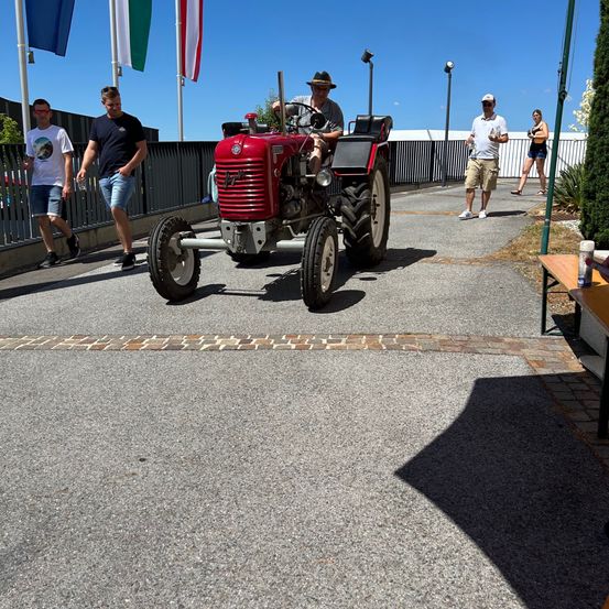 Ein Mann fährt einen roten Traktor auf einem gepflasterten Bereich. Zwei Männer gehen hinter ihm. Eine Frau geht rechts, in der Nähe eines weißen Zauns. Flaggen sind im Hintergrund zu sehen.