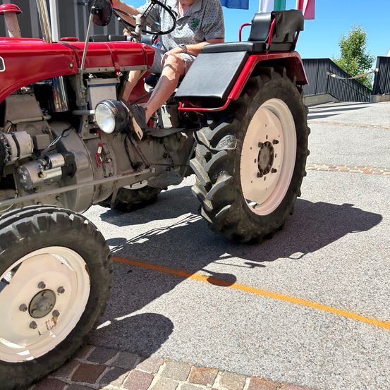 Eine Person fährt einen roten Traktor mit schwarzem Sitz auf einer asphaltierten Oberfläche. Der Schatten des Traktors fällt auf den Boden. Der Traktor hat einen großen Reifen und ein schwarzes Rad.