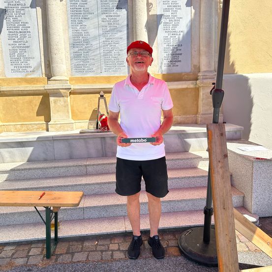 Ein älterer Mann steht auf einem gepflasterten Gehweg und hält eine grüne Metallplakette mit dem Wort 'metabo'. Er trägt einen roten Hut und eine Brille. Hinter ihm befindet sich ein Denkmal mit Namen und einer Treppe. Eine hölzerne Bank befindet sich auf der linken Seite.