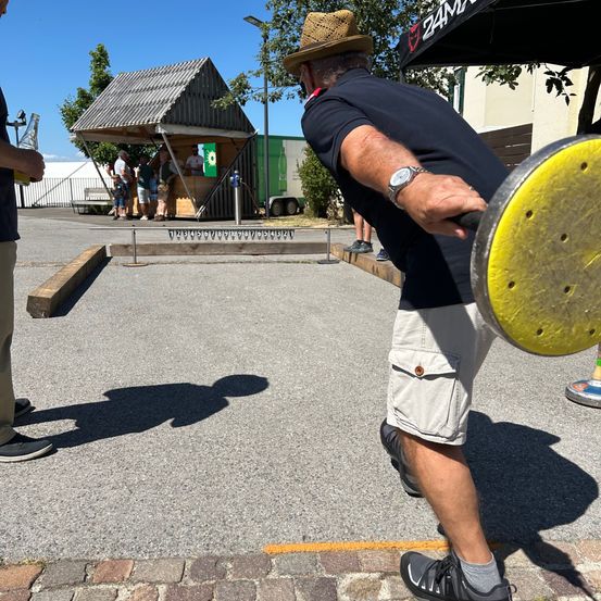 Ein Mann wirft einen gelben Scheiben auf einem Bocce-Ball-Platz, während Menschen im Hintergrund zusehen.