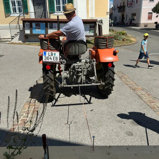 Ein Mann fährt einen orangefarbenen Traktor mit dem Nummernschild LB 364 EB auf einer kleinen asphaltierten Straße. Ein Kind geht in der Nähe. Hinter dem Traktor befinden sich Gebäude mit Fenstern, von denen eines eine Treppe hat.