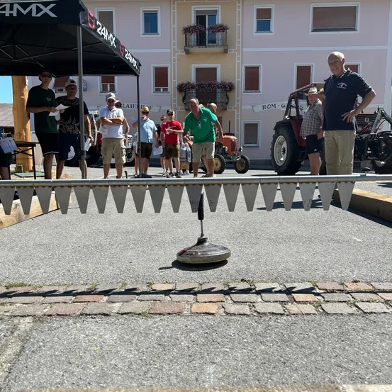 Mehrere Menschen stehen in einer Reihe und beobachten ein Spiel mit einem Gerät auf dem Boden, in der Nähe eines Zeltes und eines Gebäudes mit Fenstern und Traktoren, die davor geparkt sind.