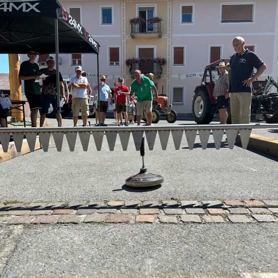 Mehrere Menschen stehen in einer Reihe und beobachten ein Spiel mit einem Gerät auf dem Boden, in der Nähe eines Zeltes und eines Gebäudes mit Fenstern und Traktoren, die davor geparkt sind.