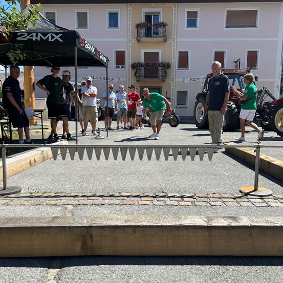Eine Gruppe von Menschen beobachtet, wie ein Mann in Grün einen Ball bei einer Sportveranstaltung wirft. Sie befinden sich auf einem Parkplatz mit einem Zelt, einem Traktor und einem Gebäude.