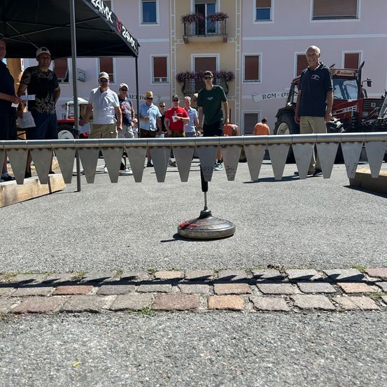 Menschen stehen um eine Bahn herum und beobachten einen Scheibenwurfwettbewerb. Ein Mann in einem grünen Shirt wirft die Scheibe. Im Hintergrund sind ein Gebäude und ein Traktor zu sehen.