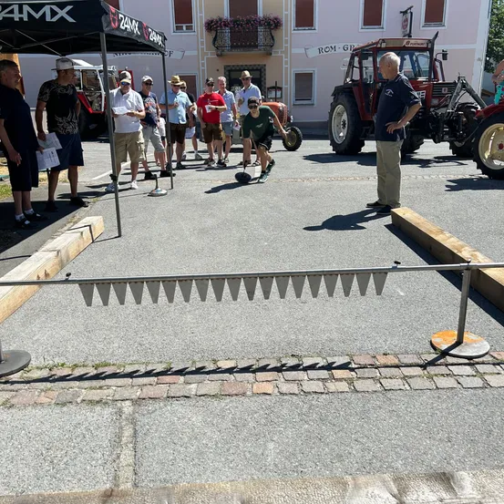 Eine Gruppe von Menschen spielt Boccia draußen auf einem Parkplatz mit einem Traktor und einem Gebäude im Hintergrund.