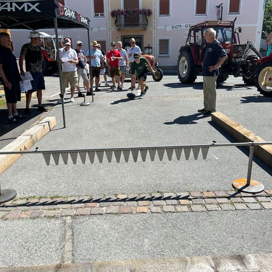 Eine Gruppe von Menschen spielt Boccia draußen auf einem Parkplatz mit einem Traktor und einem Gebäude im Hintergrund.