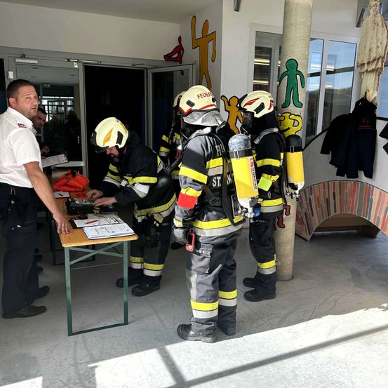 Feuerwehrleute in Schutzausrüstung stehen neben einem Tisch, einer bereitet Gerät vor. Ein Mann in einem weißen Hemd beobachtet sie.