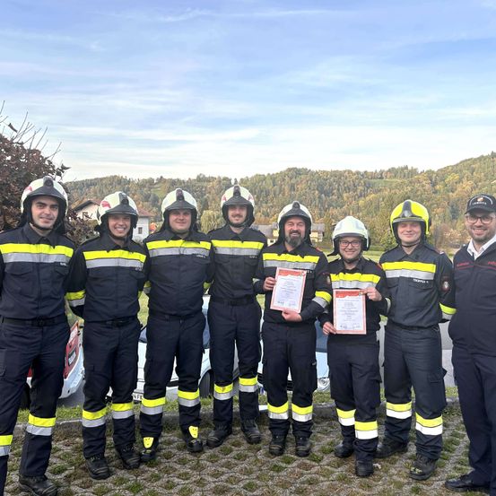 Sieben Feuerwehrleute in Uniform posieren für ein Foto, zwei halten Urkunden. Sie stehen in einer Reihe mit einem Berg und Bäumen im Hintergrund.