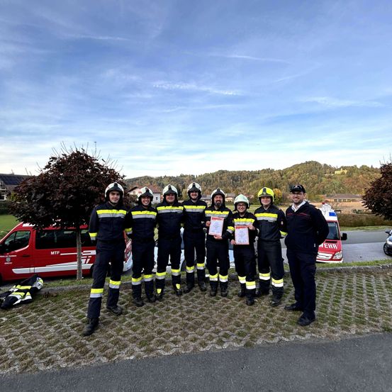 Eine Gruppe Feuerwehrleute in Uniform steht auf einem Ziegelweg und hält Urkunden in der Hand. Dahinter ist ein roter Feuerwehrwagen geparkt. Bäume und Berge sind im Hintergrund.