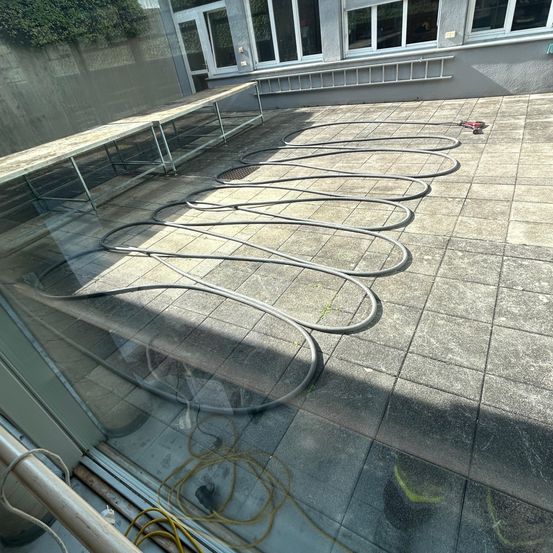 Ein Patio mit gefliestem Boden, Metalltischen, aufgewickelten Schläuchen und Kabeln, mit einem Gebäude im Hintergrund mit Fenstern und einer Leiter.