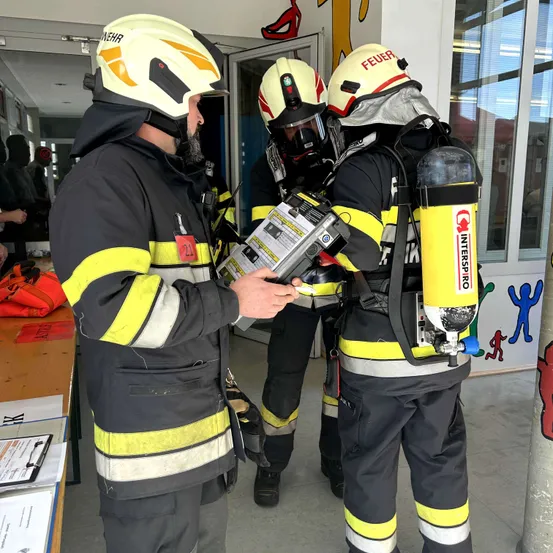 Drei Feuerwehrleute in voller Schutzausrüstung versammeln sich in einem Raum, einer von ihnen untersucht ein Gerät.