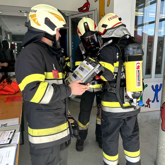 Drei Feuerwehrleute in voller Schutzausrüstung versammeln sich in einem Raum, einer von ihnen untersucht ein Gerät.