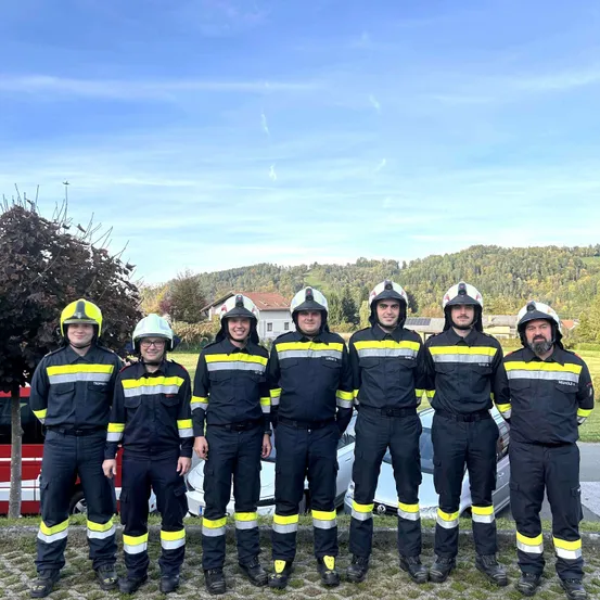 Eine Gruppe Feuerwehrleute in voller Uniform steht vor einem weißen Auto, mit einem Haus und Bergen im Hintergrund.