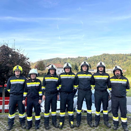 Eine Gruppe Feuerwehrleute in voller Uniform steht vor einem weißen Auto, mit einem Haus und Bergen im Hintergrund.