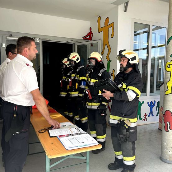 Eine Gruppe von Feuerwehrleuten steht drinnen. Ein Mann in einem weißen Hemd spricht mit ihnen. Sie tragen Helme und Masken. Papiere liegen auf einem Tisch vor ihnen.