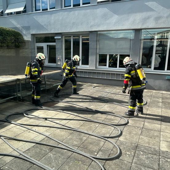 Drei Feuerwehrleute trainieren vor einem Gebäude mit Schläuchen und einem Feuerlöscher. Das Gebäude hat Glasfenster und eine graue Außenseite.