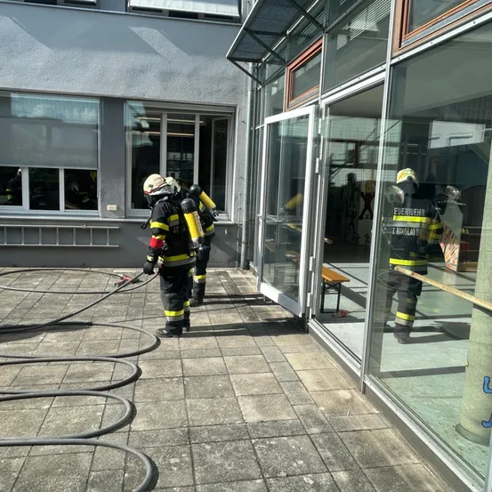 Feuerwehrleute in voller Ausrüstung sind vor einem Gebäude und sprühen Wasser. Der Eingang ist offen, und Glaswände reflektieren ihre Bilder. Ein Bank ist in der Nähe.