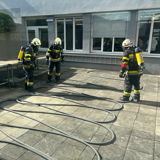 Drei Feuerwehrleute in voller Ausrüstung stehen auf einer gefliesten Außenfläche. Sie befinden sich in der Nähe mehrerer aufgewickelter Schläuche, und im Hintergrund ist ein Gebäude mit Glasfenstern zu sehen.