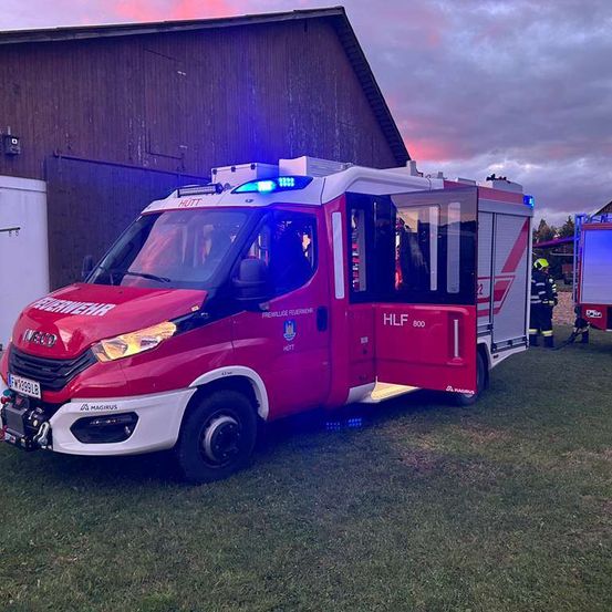 Ein rot-weißes Notfallfahrzeug mit HLF 800 auf der Seite steht auf einer Rasenfläche. Die Türen des Fahrzeugs sind geöffnet. Dahinter steht eine Person.
