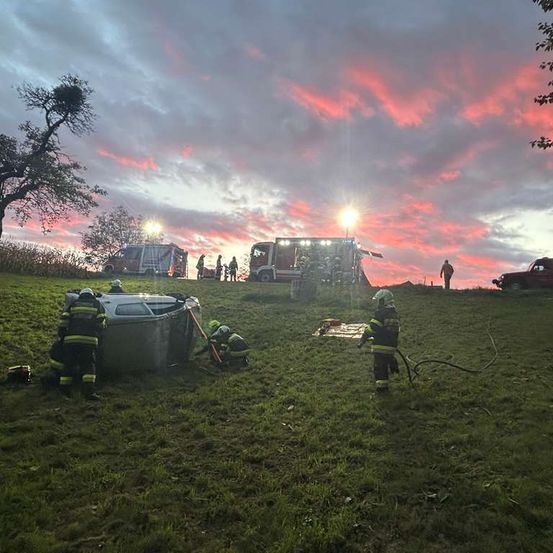 Einsatzkräfte arbeiten daran, jemanden aus einem umgekippten Fahrzeug zu befreien. Sie verwenden Werkzeuge und Seile auf einem Grasfeld. Zwei Feuerwehrwagen sind geparkt und ein Auto ist in der Nähe. Der Himmel ist eine Mischung aus Rosa und Blau.