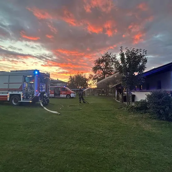 Ein Feuerwehrteam verwendet einen Schlauch, um ein Feuer in einem Gebäude unter einem farbenfrohen Sonnenuntergang zu löschen.