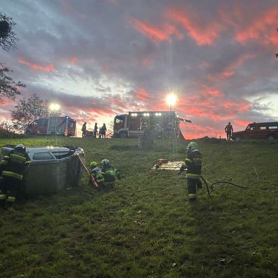 Rettungskräfte sind am Unfallort eines Fahrzeugunfalls auf einem Grashügel, mit geparkten Rettungsfahrzeugen in der Nähe. Eine Person steht rechts, und der Himmel ist vom untergehenden Sonnenlicht erhellt.