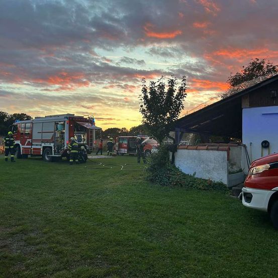 Feuerwehrleute arbeiten bei Sonnenuntergang an einem Feuerwehrauto auf einer Rasenfläche. Ein Haus steht neben ihnen.